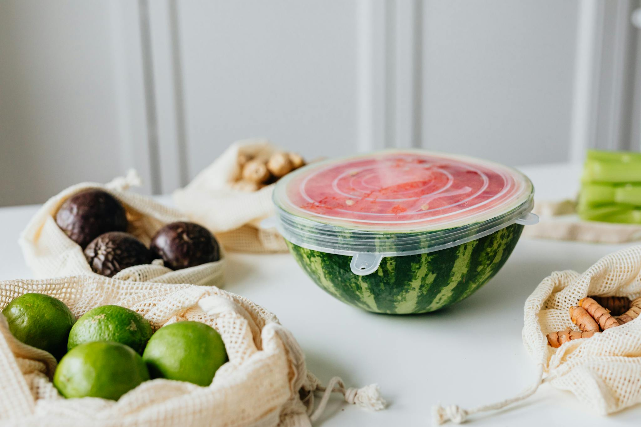 A fresh sliced watermelon with reusable food wrap, surrounded by avocados and limes on a white table.