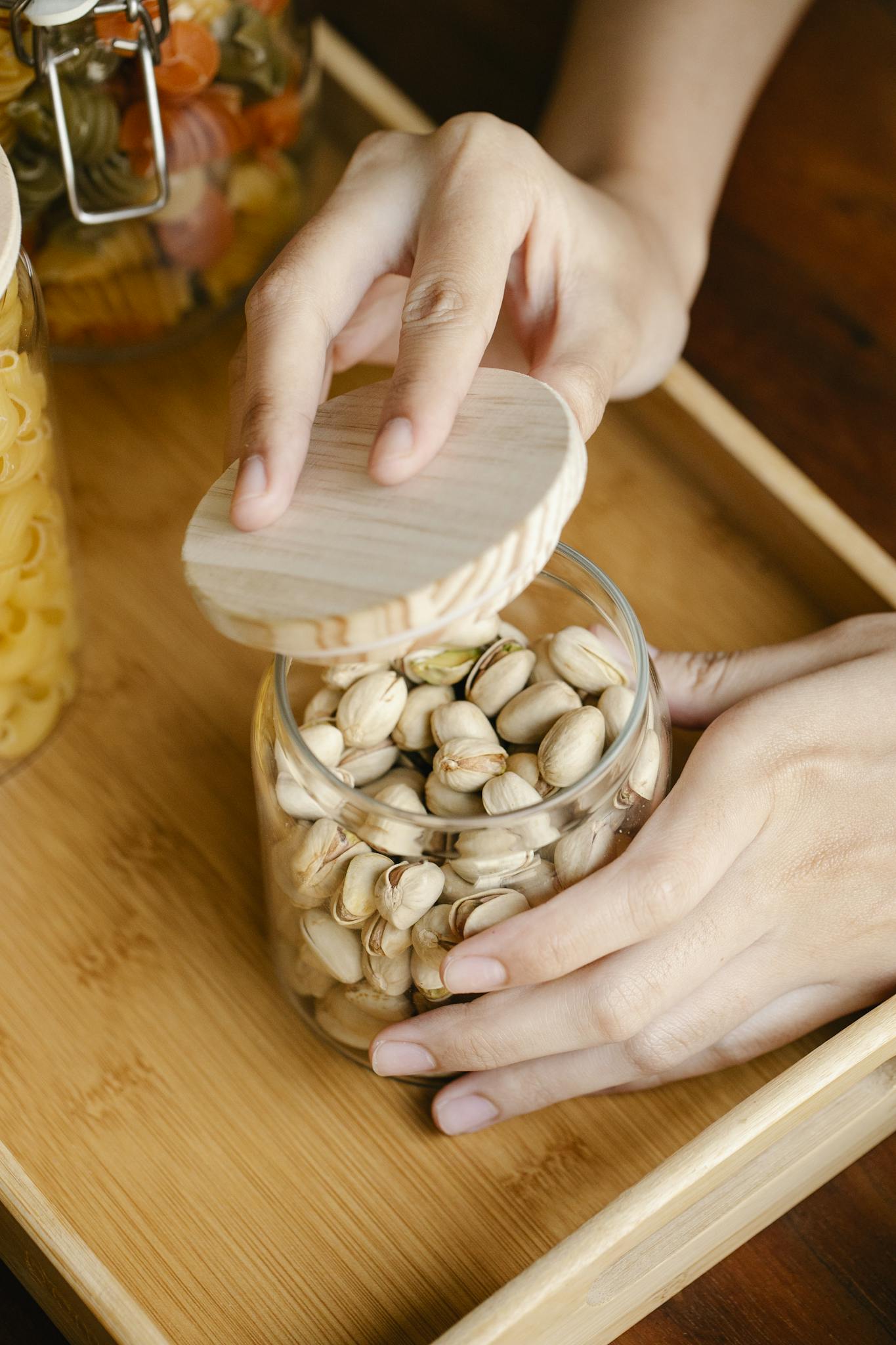 From above anonymous person opening glass jar of pistachios with wooden lid on table